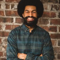 Black cisgender Man with a black afro and full black beard in front of a brick wall wearing a grey plaid shirt.
